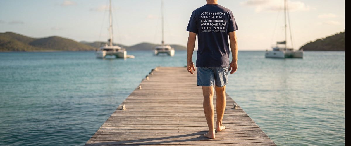 Man walking down a dock in the navy Motto Tee, back print reading Lose the phone, grab a ball, kill the engines, pour some rum, stay gone