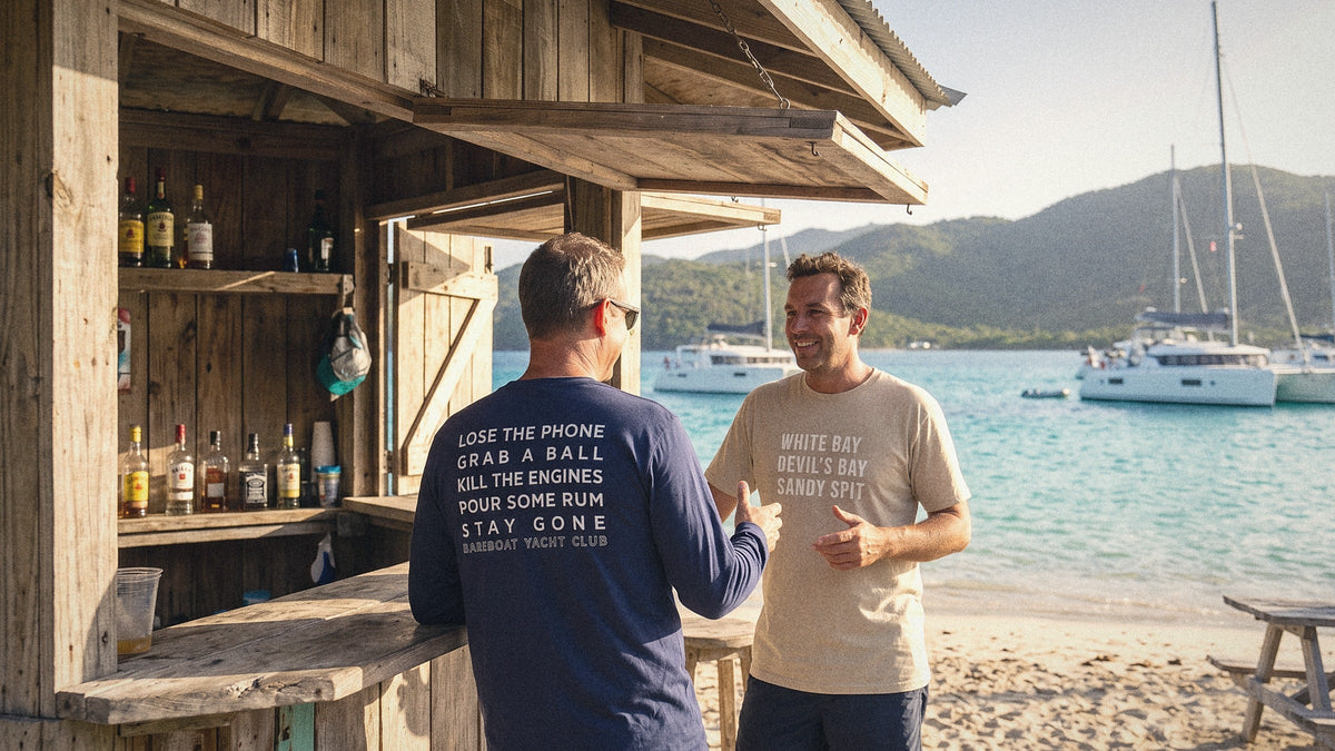 Two men talking at a beach bar in the British Virgin Islands, wearing Bareboat Yacht Club tees, catamarans at anchor behind them