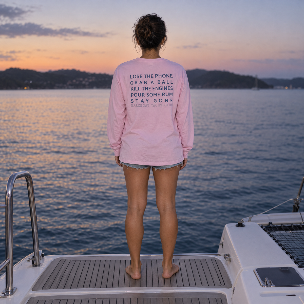 The pink Bareboat Motto Long Sleeve worn, back view: Lose the phone. Grab a ball. Kill the engines. Pour some rum. Stay gone.