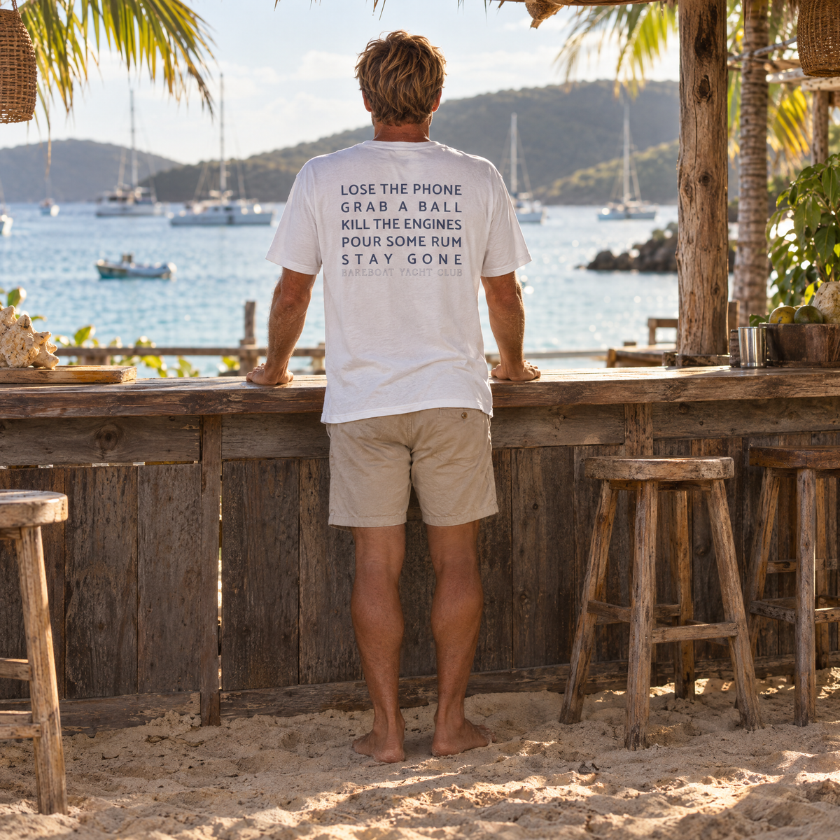 The white Bareboat Motto Tee worn, back view: Lose the phone. Grab a ball. Kill the engines. Pour some rum. Stay gone.